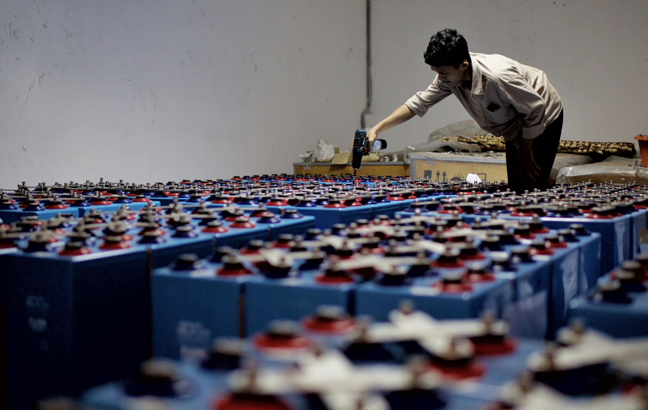 A worker checking many industrial batteries inside a facility. Indoor, industrial setting.